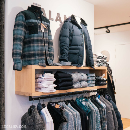 L'intérieur du magasin de vêtements pour hommes Harmony Tailor à Verviers, avec des mannequins portant des vêtements d'hiver, notamment une chemise à carreaux et une veste matelassée. Sous les mannequins, il y a une étagère en bois avec des piles de vêtements pliés, y compris des chemises blanches et des pulls. En dessous de l'étagère, des vêtements sont suspendus sur des cintres, comprenant des vestes, des chemises et des pulls.