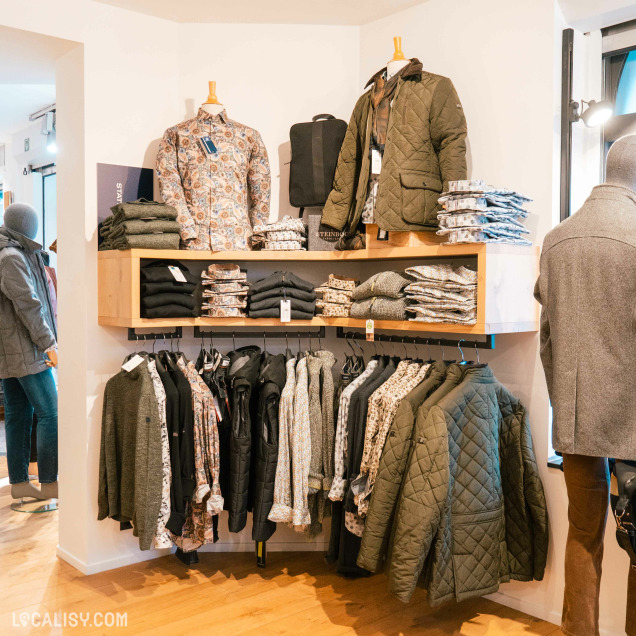 Une variété de mannequins habillés avec des vestes et des chemises, ainsi que des étagères et des cintres remplis de vêtements pliés et suspendus à l'intérieur du magasin de vêtements pour hommes Harmony Tailor à Verviers. Les vêtements incluent des vestes matelassées vertes, des chemises à motifs, et des pulls.