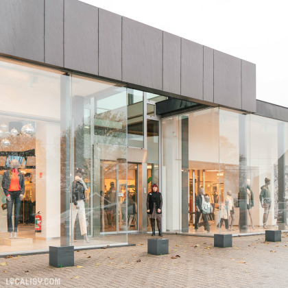 Devanture du magasin de vêtements Espace Mode à Aubel, avec de grandes vitrines en verre transparent, permettant de voir l'intérieur du magasin et les mannequins exposant des vêtements à la mode. À l'intérieur, on peut voir des clients en train de faire du shopping. Le magasin semble bien éclairé et accueillant.