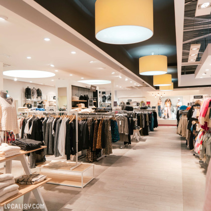 Plusieurs rangées de vêtements suspendus sur des cintres, avec des étagères contenant des vêtements pliés à l'intérieur du magasin de vêtements Espace Mode à Aube. Le magasin est décoré avec des luminaires suspendus circulaires et des mannequins habillés en arrière-plan.