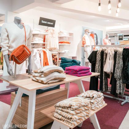 Des mannequins habillés, des étagères et des tables avec des vêtements pliés, notamment des pulls et des écharpes dans le magasin de vêtements Espace Mode à Aube. Un mannequin porte un pull blanc avec un sac orange. Il y a également des vêtements suspendus sur des cintres à droite. L'éclairage est chaleureux avec des ampoules suspendues au plafond.