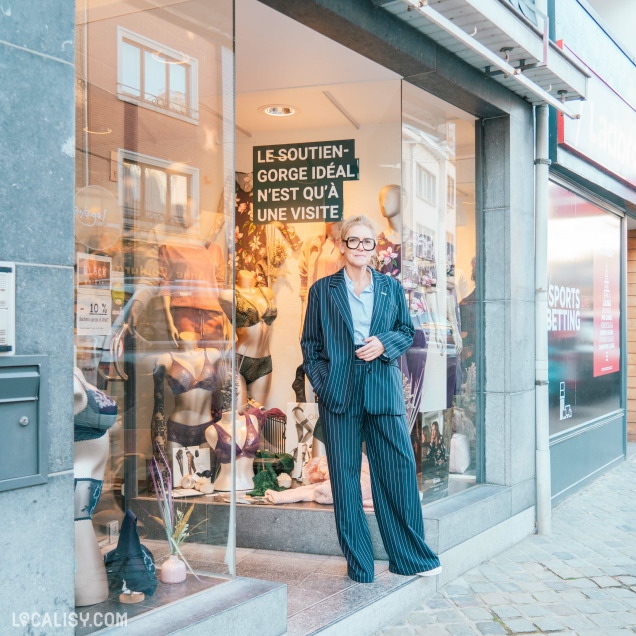 Devanture du magasin de lingerie Jardin Secret à Malmedy, avec une vitrine qui expose divers ensembles de lingerie sur des mannequins. Un individu en costume rayé se tient devant la vitrine. Une affiche à l'intérieur de la vitrine indique : LE SOUTIEN-GORGE IDÉAL N'EST QU'À UNE VISITE. À droite, on peut voir une enseigne pour les paris sportifs.