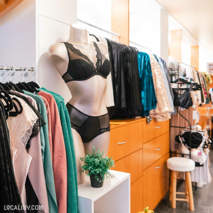 L'intérieur du magasin de lingerie Jardin Secret à Malmedy, avec des vêtements suspendus sur des cintres, des tiroirs en bois, et un mannequin portant des sous-vêtements. Il y a également une petite plante verte sur une étagère blanche. Les vêtements exposés incluent des chemises, des vestes, et des articles de lingerie.