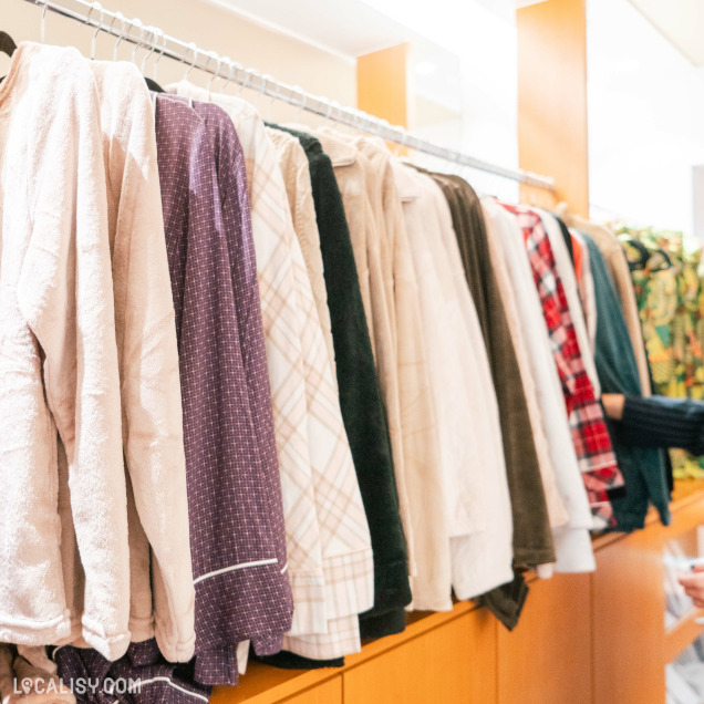 Une rangée de vêtements suspendus sur des cintres dans le magasin de lingerie Jardin Secret à Malmedy. Les vêtements sont principalement des chemises et des vestes de différentes couleurs et motifs, allant du beige au violet, avec des motifs à carreaux et unis. L'image est lumineuse et bien éclairée, mettant en valeur les vêtements exposés.