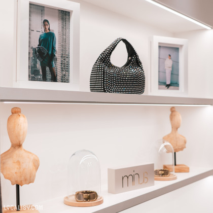 Une étagère d'exposition dans le magasin de vêtements Habit&Vous à Tiff. Sur l'étagère supérieure, il y a deux cadres photo avec des images de personnes, et entre les deux, un sac à main noir avec des motifs blancs. Sur l'étagère inférieure, il y a deux bustes en bois, deux cloches en verre contenant des bracelets, et un bloc avec le texte minus.