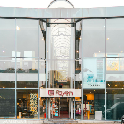 Devanture du magasin d'ameublement et de décoration maison Fayen à Verviers. Le magasin est situé dans un bâtiment moderne avec de grandes vitres. On peut voir l'entrée principale avec une enseigne lumineuse indiquant Fayen. À l'intérieur, il y a des escaliers menant à un étage supérieur. À droite de l'entrée, il y a une vitrine avec le nom Tollens et des produits exposés.