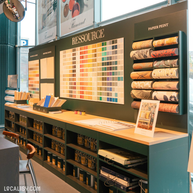 Un mur du magasin d'ameublement et de décoration maison Fayen à Verviers, avec des échantillons de couleurs de peinture et de papier peint. Le mur est divisé en plusieurs sections, dont une grande section centrale intitulée RESSOURCE avec une large gamme de couleurs. À droite, il y a une section intitulée PAPIER PEINT avec des rouleaux de papier peint de différents motifs. En dessous, il y a des étagères contenant des pots de peinture et des classeurs. Des échantillons de bois et des brochures sont disposés sur le comptoir.