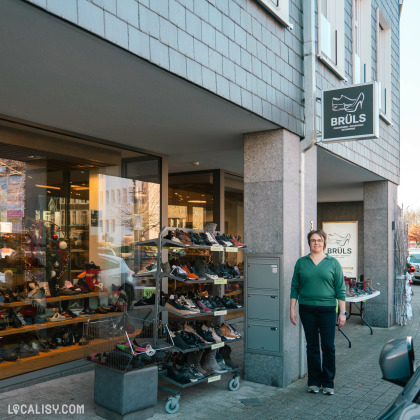 Devanture du magasin de chaussures Maison Brüls à Malmedy. On peut voir une vitrine avec plusieurs étagères de chaussures exposées à l'extérieur du magasin. Il y a également un panneau avec le logo du magasin et le nom "Brüls" affiché au-dessus de l'entrée. Le magasin semble être situé dans un bâtiment moderne avec une façade en pierre et des fenêtres vitrées.