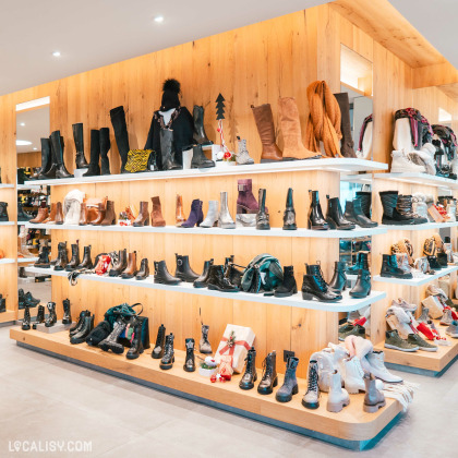 Des étagères en bois remplies de diverses bottes et chaussures, organisées par style et couleur dans le magasin de chaussures Maison Brüls à Malmedy. Il y a des bottes hautes, des bottines et des chaussures de différentes matières et couleurs, allant du noir au marron, en passant par des teintes plus claires et métalliques.