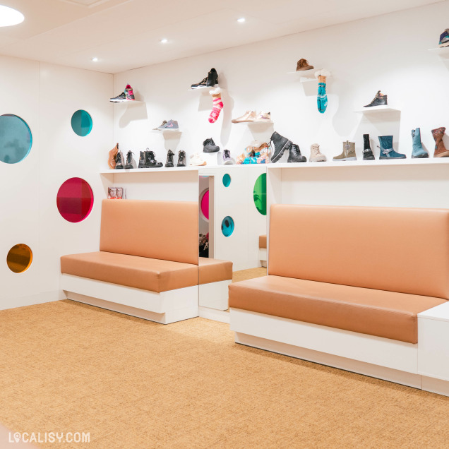 Des chaussures, probablement destiné aux enfants, avec des bancs rembourrés de couleur beige clair et des étagères blanches sur lesquelles sont exposées diverses chaussures dans le magasin de chaussures Maison Brüls à Malmedy. Les murs sont décorés de cercles colorés transparents, ajoutant une touche ludique à l'espace. Le sol est recouvert d'un tapis beige. Le magasin semble bien éclairé avec des spots au plafond.