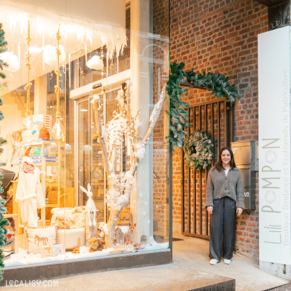 Devanture du magasin de puériculture Lili Pompon à Visé. La vitrine est décorée de manière festive avec des éléments hivernaux comme des branches enneigées, des guirlandes lumineuses et des décorations de Noël. À l'intérieur, on peut voir des vêtements et des jouets pour enfants. Une personne se tient à l'extérieur du magasin, à côté d'une couronne de Noël accrochée à un mur en briques. Le texte sur la vitrine indique Boutique Tendance et Ecofriendly de Puériculture.