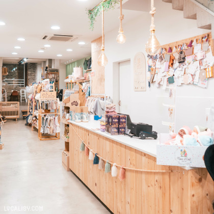 L'intérieur du magasin de puériculture Lili Pompon à Visé, avec des installations en bois et un design moderne et épuré. Le comptoir est décoré avec une guirlande de pompons colorés, et divers articles sont exposés sur et derrière le comptoir, y compris des boîtes et une caisse enregistreuse. Les murs derrière le comptoir sont décorés de nombreuses notes, cartes et petits objets épinglés, ajoutant une touche personnelle et chaleureuse à l'espace. Les étagères et les portants à travers le magasin sont soigneusement garnis de vêtements, jouets et autres marchandises.