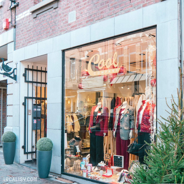 Devanture du magasin de vêtements pour femmes Cool Company à Visé. La vitrine présente plusieurs mannequins habillés avec des vêtements élégants, principalement dans des tons de rouge et de gris. À l'intérieur de la vitrine, on peut voir divers accessoires et articles de mode exposés. L'extérieur du magasin est en briques avec des éléments de décoration modernes, y compris deux grandes jardinières avec des plantes sphériques.