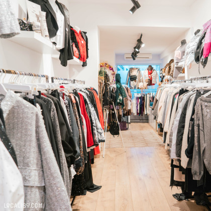 L'intérieur du magasin de vêtements pour femmes Cool Company à Visé, avec des rangées de vêtements suspendus sur des cintres de chaque côté de l'allée centrale. Les vêtements incluent des vestes, des manteaux, des pulls et d'autres articles de mode. Le sol est en bois clair, et il y a des spots lumineux au plafond pour éclairer l'espace.