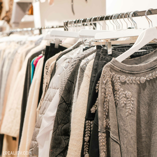 Une rangée de vêtements suspendus sur des cintres dans le magasin de vêtements pour femmes Cool Company à Visé. Les vêtements incluent des pulls, des vestes et des jupes, principalement dans des tons neutres comme le gris, le blanc et le noir. Certains vêtements sont ornés de perles ou de broderies, ajoutant une touche d'élégance.