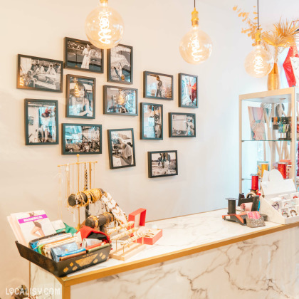 Un comptoir en marbre avec divers articles exposés, tels que des bracelets, des porte-monnaie et des boîtes dans le magasin de vêtements pour femmes Cool Company à Visé. Au-dessus du comptoir, il y a plusieurs cadres photo accrochés au mur, contenant des images en noir et blanc. Des lampes suspendues avec des ampoules décoratives éclairent la scène. À droite, une vitrine contient d'autres produits.