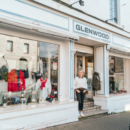 Devanture du magasin de vêtements pour hommes Glenwood à Spa. La vitrine expose divers vêtements, dont un manteau rouge et un pull blanc avec une écharpe rouge. Une personne se tient à l'entrée du magasin, portant un pull blanc et un pantalon noir. L'affichage est soigné et attrayant, ce qui peut attirer les clients.