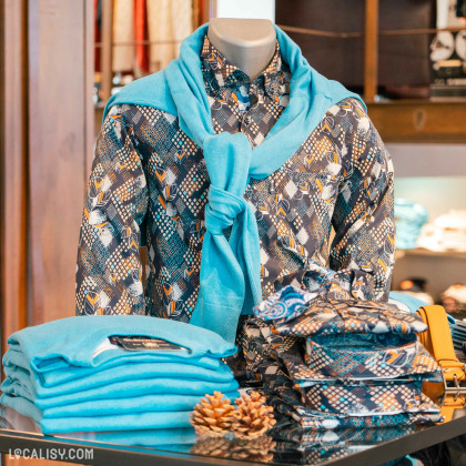 Un présentoir dans le magasin de vêtements pour hommes Glenwood à Spa, mettant en avant une chemise à motifs géométriques accompagnée d'un pull bleu clair noué sur les épaules. Sur la table, des vêtements soigneusement pliés et assortis complètent l'agencement, reflétant une ambiance soignée et moderne. Des éléments décoratifs comme des pommes de pin ajoutent une touche chaleureuse.