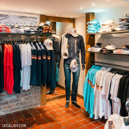 L'intérieur du magasin de vêtements pour hommes Glenwood à Spa, avec des vêtements et accessoires liés à l'univers automobile. On y voit des chemises, polos et casquettes ornés de logos et motifs en rapport avec Spa-Francorchamps, célèbre circuit de course. Les articles, soigneusement organisés sur des étagères et portants, reflètent un style sportif et élégant, ciblant les amateurs de courses automobiles et de mode décontractée.