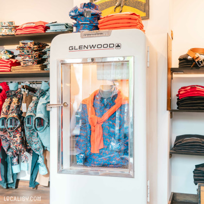 Une vitrine avec un mannequin habillé d'une chemise bleue à motifs paisley et un pull orange noué autour du cou dans le magasin de vêtements pour hommes Glenwood à Spa. La vitrine porte l'inscription GLENWOOD. Autour de la vitrine, des étagères contiennent des vêtements pliés, notamment des chemises colorées et des pantalons. Le magasin semble bien organisé et met en avant des vêtements aux motifs et couleurs vives.