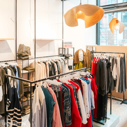 L'intérieur du magasin d'accessoires de mode Côté Rue à Heusy, avec plusieurs portants avec des vêtements suspendus, incluant des chemises, des pulls, des robes et des vestes de différentes couleurs et styles. On peut également voir des étagères avec des vêtements pliés. Deux grandes lampes suspendues au plafond ajoutent une touche décorative à l'espace.