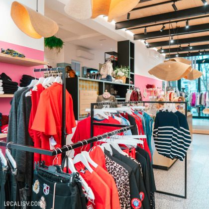 L'intérieur du magasin d'accessoires de mode Côté Rue à Heusy, avec des luminaires suspendus en forme de chapeaux. Les vêtements sont disposés sur des portants et des étagères, avec une variété de couleurs et de styles, notamment des chemises rouges, des pulls rayés et des vêtements à motifs léopard. Le magasin a également des éléments de décoration comme des plantes et des étagères avec des accessoires.