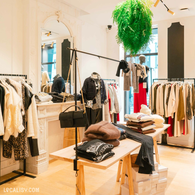 Plusieurs portants remplis de vêtements divers, tels que des manteaux, des pulls, et des pantalons dans le magasin d'accessoires de mode Côté Rue à Heusy. Au centre, il y a une table en bois avec des vêtements pliés, notamment des pulls et des jeans. Un grand miroir ornée est visible à l'arrière, reflétant une partie du magasin. Une plante verte suspendue ajoute une touche de verdure à l'espace.