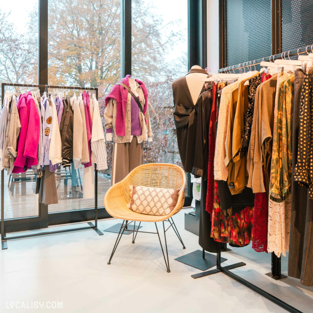 Des portants remplis de vêtements de différentes couleurs et styles, allant des vestes et chemises aux robes et pulls dans le magasin d'accessoires de mode Côté Rue à Heusy. Un mannequin porte une tenue composée d'un pantalon large, d'une chemise et d'un pull violet noué autour des épaules. Un fauteuil en osier avec un coussin est placé au centre de la pièce, offrant un espace confortable pour s'asseoir.