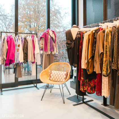 Des portants remplis de vêtements de différentes couleurs et styles, allant des vestes et chemises aux robes et pulls dans le magasin d'accessoires de mode Côté Rue à Heusy. Un mannequin porte une tenue composée d'un pantalon large, d'une chemise et d'un pull violet noué autour des épaules. Un fauteuil en osier avec un coussin est placé au centre de la pièce, offrant un espace confortable pour s'asseoir.