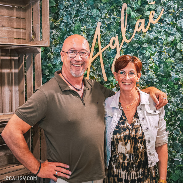 Deux personnes souriantes devant un mur de feuilles vertes, avec l'enseigne Aglaé écrite en lettres dorées. Le décor semble chaleureux et naturel, évoquant une ambiance conviviale dans le magasin de vêtements pour femmes Aglaé à Malmedy.