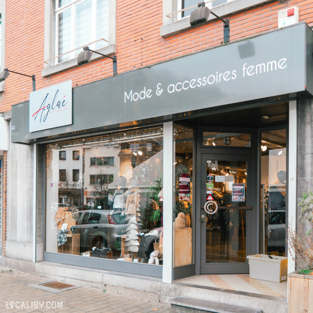 Devanture du magasin de vêtements pour femmes Aglaé à Malmedy. Le magasin est spécialisé dans la mode et les accessoires pour femmes, comme indiqué par l'enseigne Mode & accessoires femme. La vitrine est en verre, permettant de voir l'intérieur du magasin où sont exposés divers articles de mode. L'enseigne du magasin est grise avec le nom Aglaé écrit en lettres cursives.