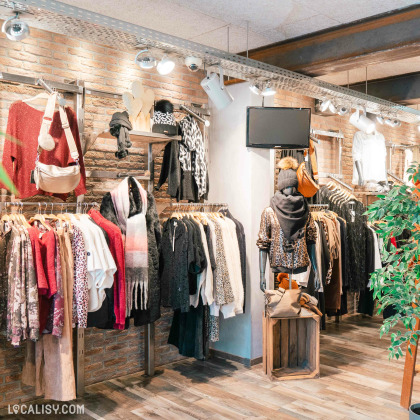 L'intérieur du magasin de vêtements pour femmes Aglaé à Malmedy, avec des murs en briques apparentes et les vêtements suspendus sur des cintres le long des murs. Il y a une variété de vêtements, y compris des pulls, des vestes, des écharpes et des sacs à main. Un mannequin est également visible, habillé avec des vêtements à motifs léopard et portant un bonnet avec un pompon.