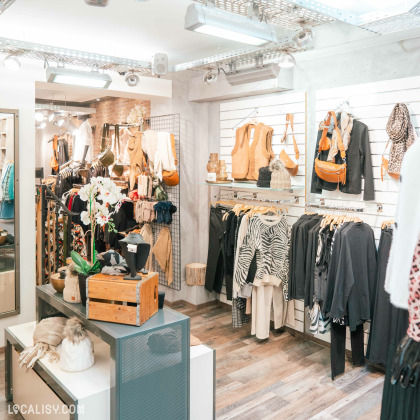 Une variété de vêtements, sacs à main, écharpes et autres accessoires disposés de manière organisée sur des étagères et des portants dans le magasin de vêtements pour femmes Aglaé à Malmedy. Il y a également des éléments de décoration comme des plantes et des mannequins.