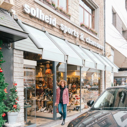 Devanture du magasin de chaussures Solheid Chaussures à Malmedy. La vitrine du magasin expose divers articles, principalement des chaussures et des sacs à main. Une personne est en train de sortir du magasin. La devanture est décorée avec des plantes et des lumières, et il y a un arbre de Noël décoré à gauche de l'image. Une voiture est garée devant le magasin.