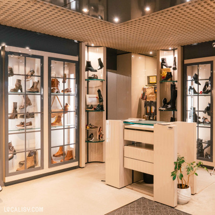 L'intérieur du magasin de chaussures Solheid Chaussures à Malmedy, avec plusieurs étagères vitrées et éclairées exposant divers modèles de chaussures, allant des bottes aux chaussures de ville. Au centre, il y a un comptoir de réception avec des tiroirs et une plante en pot à côté. Le plafond est décoré de panneaux en treillis, et l'éclairage est doux et chaleureux, créant une ambiance accueillante.