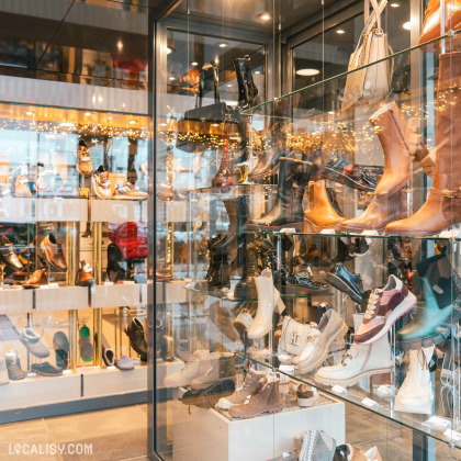 Une vitrine bien éclairée et présente une variété de chaussures, y compris des bottes, des baskets et des chaussures habillées dans le magasin de chaussures Solheid Chaussures à Malmedy. Les chaussures sont disposées sur plusieurs étagères en verre, ce qui permet de bien les voir.