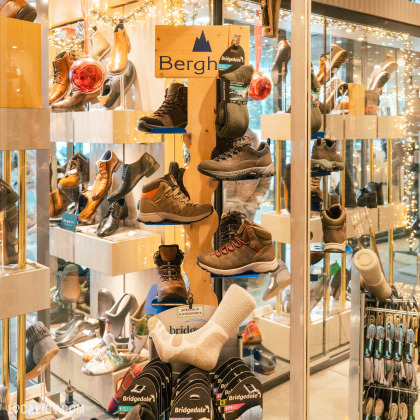 Plusieurs paires de chaussures de randonnée exposées sur des étagères et des présentoirs, ainsi que des chaussettes de la marque Bridgedale dans le magasin de chaussures Solheid Chaussures à Malmedy. La vitrine est décorée avec des lumières et des boules de Noël, ce qui suggère une ambiance festive.