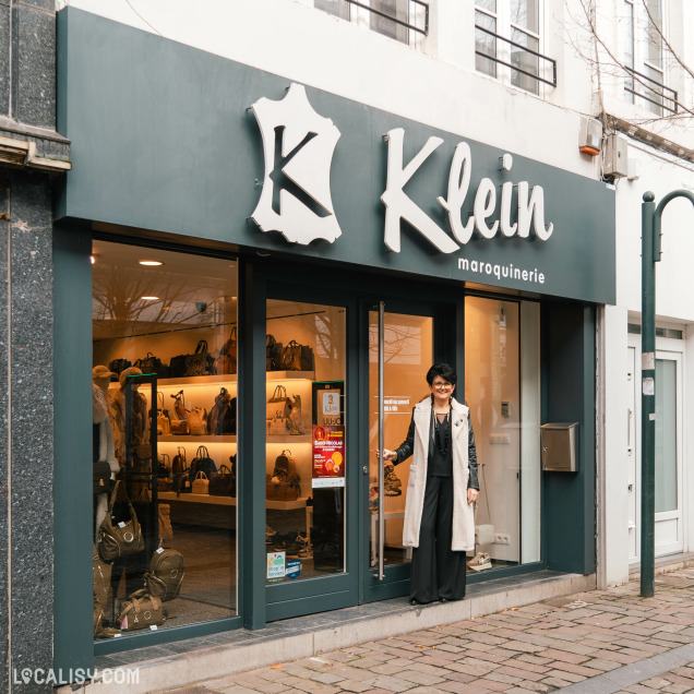 Devanture du magasin de maroquinerie Maroquinerie Klein à Verviers. La façade est de couleur sombre avec une enseigne blanche affichant le nom du magasin et un logo en forme de peau de cuir. À travers les vitrines, on peut voir des étagères illuminées exposant divers sacs à main et articles de maroquinerie. Une personne se tient à l'entrée du magasin, tenant la porte ouverte. Le trottoir devant le magasin est pavé.