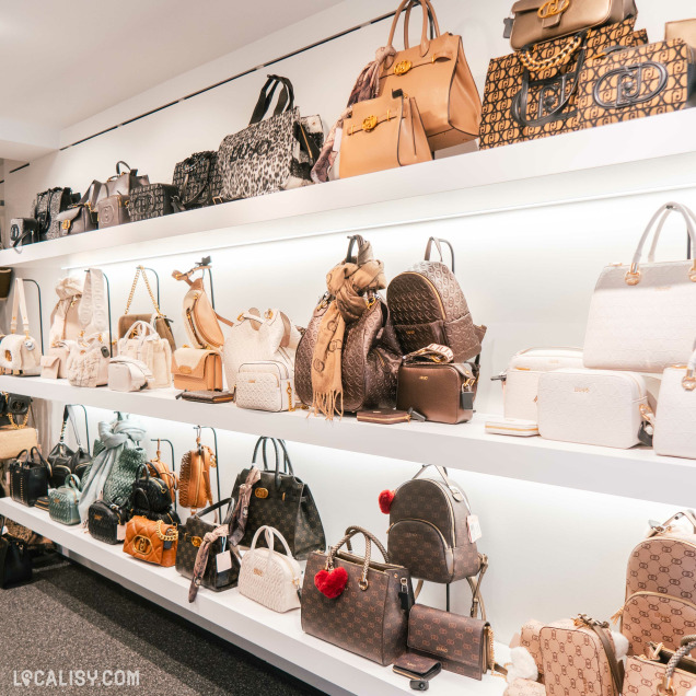 Des étagères remplies de divers sacs à main de différentes tailles, couleurs et marques dans le magasin de maroquinerie Maroquinerie Klein à Verviers. Les sacs sont soigneusement disposés sur trois niveaux d'étagères, avec une variété de styles allant des sacs à dos aux sacs à main et aux pochettes. Les couleurs dominantes sont le beige, le marron et le blanc, avec quelques touches de noir et de motifs imprimés.