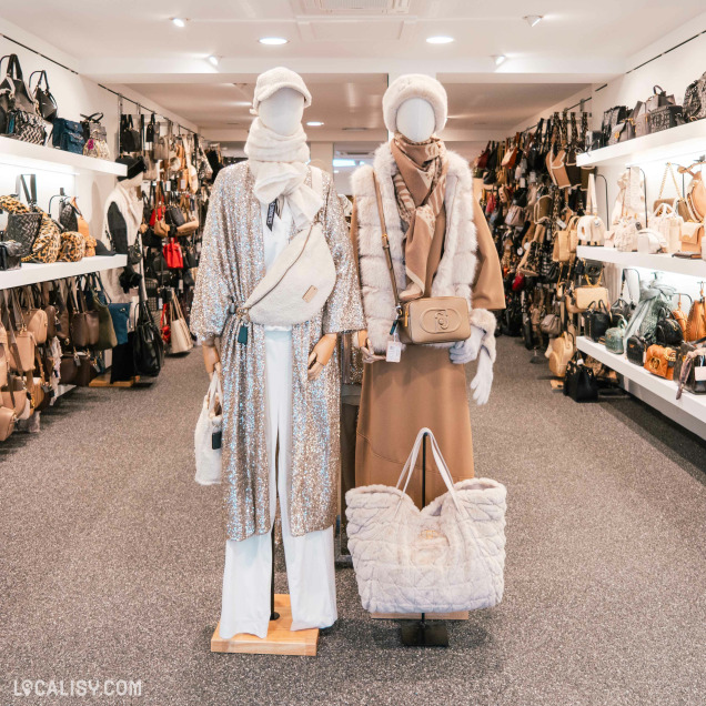 L'intérieur du magasin de maroquinerie Maroquinerie Klein à Verviers, spécialisé dans la vente de sacs à main et d'accessoires de mode, avec deux mannequins placés au centre, habillés de manière élégante et hivernale, avec des manteaux, des écharpes et des sacs en peluche. Les étagères autour des mannequins sont remplies de divers sacs à main de différentes tailles, couleurs et styles.