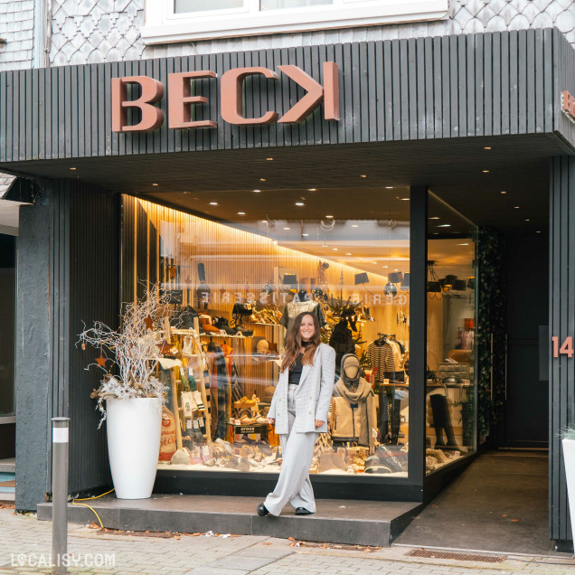 Devanture du magasin de chaussures Beck by Charl à Malmedy. La façade est moderne avec des lettres en relief formant le nom BECK en haut. La vitrine est bien éclairée et présente divers articles de mode, notamment des vêtements, des accessoires et des chaussures. Devant le magasin, une personne est debout, habillée de manière élégante. Un grand pot blanc avec des branches décoratives se trouve à gauche de l'entrée.