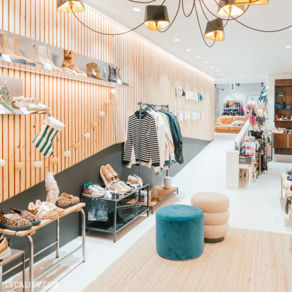 Une décoration moderne avec des murs en bois et des luminaires suspendus à l'intérieur du magasin de chaussures Beck by Charl à Malmedy. À gauche, des étagères et des bancs exposent divers types de chaussures, tandis qu'un portant présente des vêtements, notamment des pulls et des vestes. Au centre, il y a des tabourets ronds et un tapis beige.