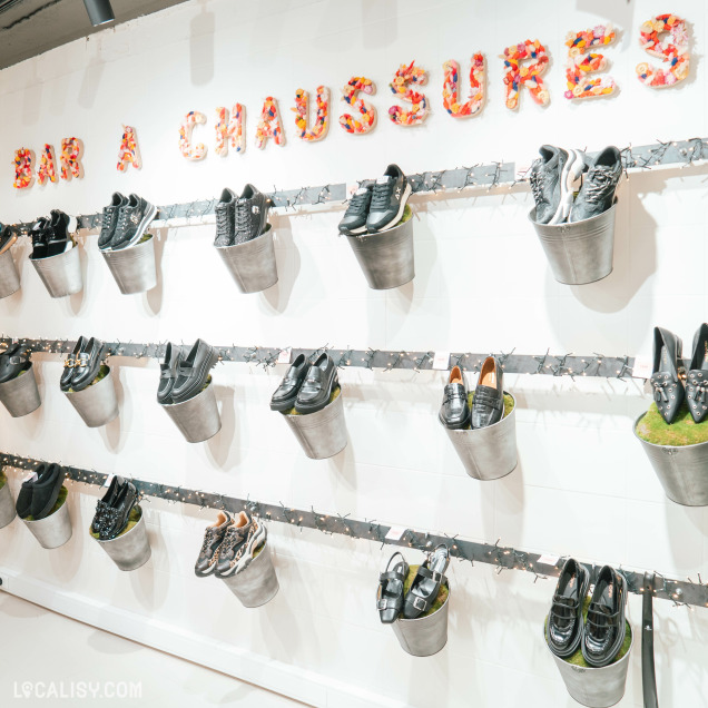 Un mur d'exposition de chaussures dans le magasin de chaussures Beck by Charl à Malmedy. Les chaussures sont placées dans des seaux métalliques fixés au mur en deux rangées horizontales. Au-dessus des chaussures, il y a une inscription en lettres colorées qui dit BAR A CHAUSSURES. Les seaux contiennent différents types de chaussures, principalement des baskets et des chaussures habillées.