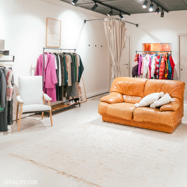 Un canapé en cuir marron avec des coussins blancs, un tapis blanc moelleux, et une chaise blanche à accoudoirs en bois dans le magasin de chaussures Beck by Charl à Malmedy. À gauche, il y a un portant avec divers vêtements, dont un costume rose vif. À droite, un autre portant contient des vêtements colorés. Il y a également un rideau beige suspendu, probablement pour une cabine d'essayage.