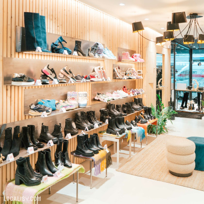 Des murs en bois clair avec des étagères en métal où sont exposées diverses chaussures dans le magasin de chaussures Beck by Charl à Malmedy. Les étagères du haut présentent principalement des baskets et des chaussures de sport colorées, tandis que les étagères du bas sont remplies de bottes noires de différents styles. Le magasin est bien éclairé avec des luminaires modernes suspendus au plafond.