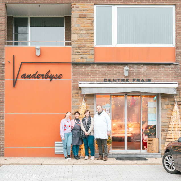 Devanture de la Boucherie Charcuterie Vanderbyse à Villers-L'Evêque. On y voit cinq personnes debout devant l'entrée du magasin, qui est décorée avec des lumières et des décorations de Noël. Le bâtiment est en briques avec une partie peinte en orange. Le nom Vanderbyse est écrit en lettres cursives sur le mur orange à gauche, tandis que CENTRE FRAIS est inscrit au-dessus de l'entrée principale.