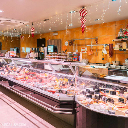 Un comptoir réfrigéré rempli de divers produits de charcuterie et de viande, étiquetés avec des petites pancartes dans la Boucherie Charcuterie Vanderbyse à Villers-L'Evêque. Au-dessus du comptoir, des décorations de Noël et des guirlandes lumineuses sont suspendues. À l'arrière, des étagères contiennent des bouteilles de vin, des pots de confiture et d'autres produits. Des saucissons et des jambons sont accrochés au mur.