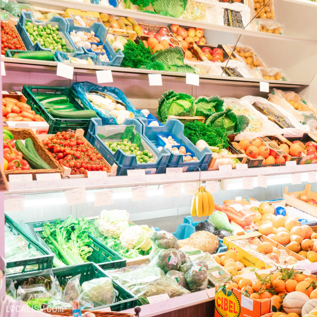 Une étagère de fruits et légumes dans la Boucherie Charcuterie Vanderbyse à Villers-L'Evêque. On y voit une variété de produits frais, notamment des tomates, des concombres, des poivrons, des choux, des pommes de terre, des oranges, des bananes, et d'autres légumes verts. Les produits sont disposés dans des caisses en plastique et des paniers, avec des étiquettes de prix visibles au-dessus de chaque section.