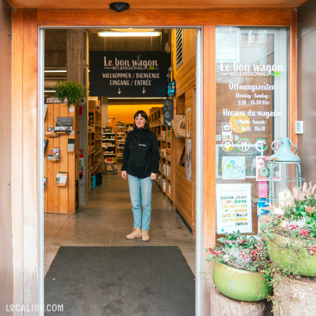 Devanture du magasin d'alimentation naturelle Le Bon Wagon à Eupen. Une personne se tient à l'entrée du magasin, qui est bien éclairé et organisé avec des étagères remplies de produits. À gauche de l'entrée, un panneau en bois affiche des brochures et des informations. Sur la porte vitrée à droite, les heures d'ouverture du magasin sont indiquées en allemand et en français : Dienstag - Samstag: 9.30 - 18.30 Uhr et Lundi - Samedi: 9h30 - 18h30. Des décorations florales embellissent l'extérieur de l'entrée.