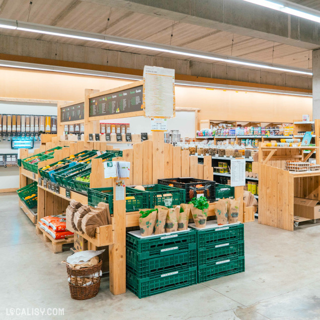 L'intérieur du magasin d'alimentation naturelle Le Bon Wagon à Eupen. Le magasin est bien organisé avec des étagères en bois contenant des caisses vertes remplies de divers produits, principalement des fruits et légumes. Il y a également des sacs en papier et des paniers en osier pour les clients. À l'arrière-plan, on peut voir des distributeurs en vrac et des étagères avec d'autres produits alimentaires.